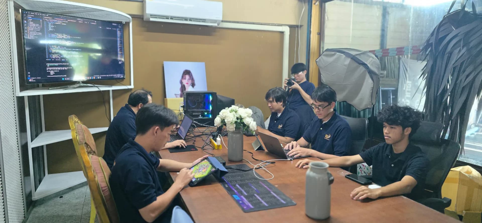 ภาพถ่ายบรรยากาศการทำงานจริง (Behind the Scenes) ของทีมงาน BizNewEra ในห้องทำงาน (Back Office) ที่เต็มไปด้วยความกระตือรือร้น โดยกลุ่มคนรุ่นใหม่ในทีมสวมเสื้อโปโลสีน้ำเงินเข้ม (Navy) ที่มีโลโก้บริษัท กำลังนั่งล้อมโต๊ะทำงานตัวยาวเพื่อปฏิบัติภารกิจต่าง ๆ ร่วมกันอย่างตั้งใจ  ในภาพมีทีมงานฝ่ายต่าง ๆ ทั้งนักพัฒนาที่กำลังเช็กโค้ดบนหน้าจอใหญ่และโน้ตบุ๊ก, ฝ่ายคอนเทนต์และกลยุทธ์ที่กำลังทำงานผ่านแท็บเล็ตและอุปกรณ์ดิจิทัล, รวมถึงการจดบันทึกและเตรียมข้อมูล โดยรอบโต๊ะเต็มไปด้วยอุปกรณ์ทำงานอย่าง แล็ปท็อป สายเชื่อมต่อ ปลั๊กไฟ และแก้วน้ำ แสดงถึงการทำงานที่เป็นระบบและการร่วมแรงร่วมใจของทีมงานหลังบ้านในการขับเคลื่อนแบรนด์และพัฒนาธุรกิจในยุคดิจิทัล